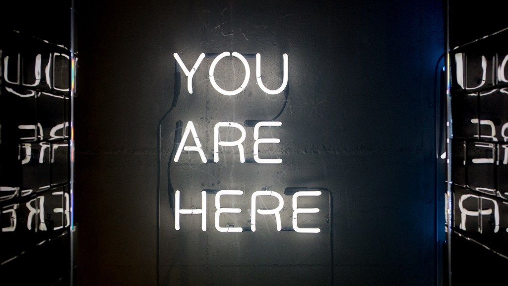 White neon sign reading "You Are Here" hanging on a black wall
