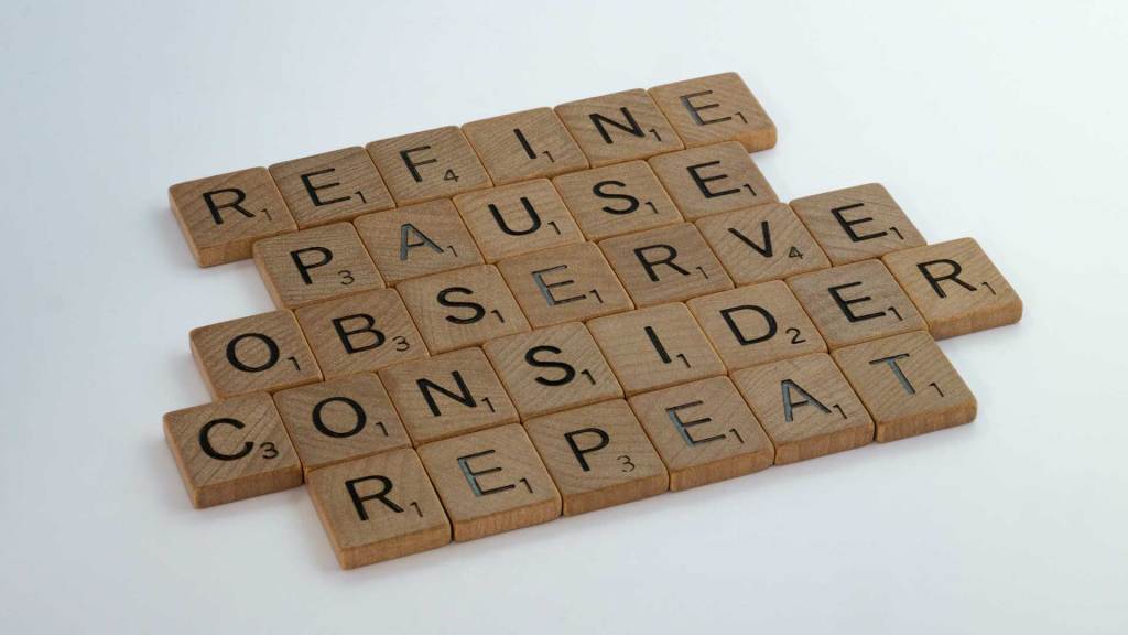 Wooden Scrabble tiles spelling out Refine Pause Observe Consider Repeat against a white backdrop.