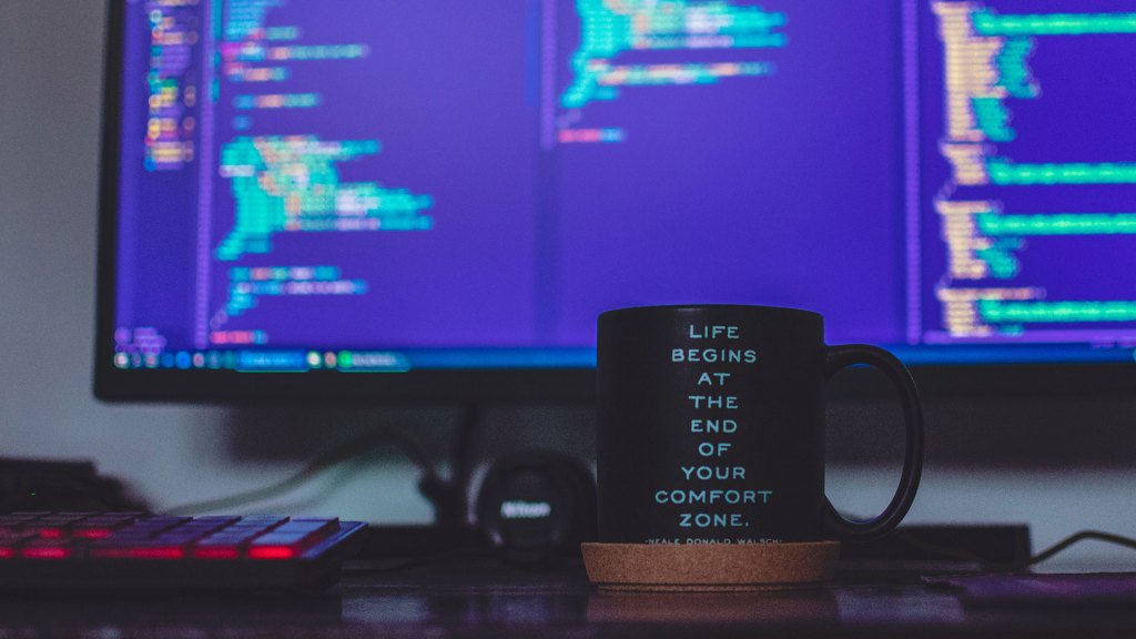 Black coffee mug with "Life begins at the end of your comfort zone" written on it sitting in front of computer screen with code on it.