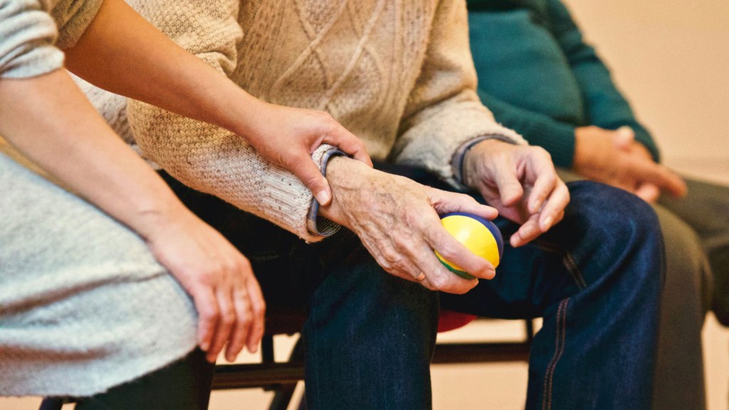 Three people site in a row. An older man holds a small ball in his hand while another person places their hand gently on his wrist in comfort.